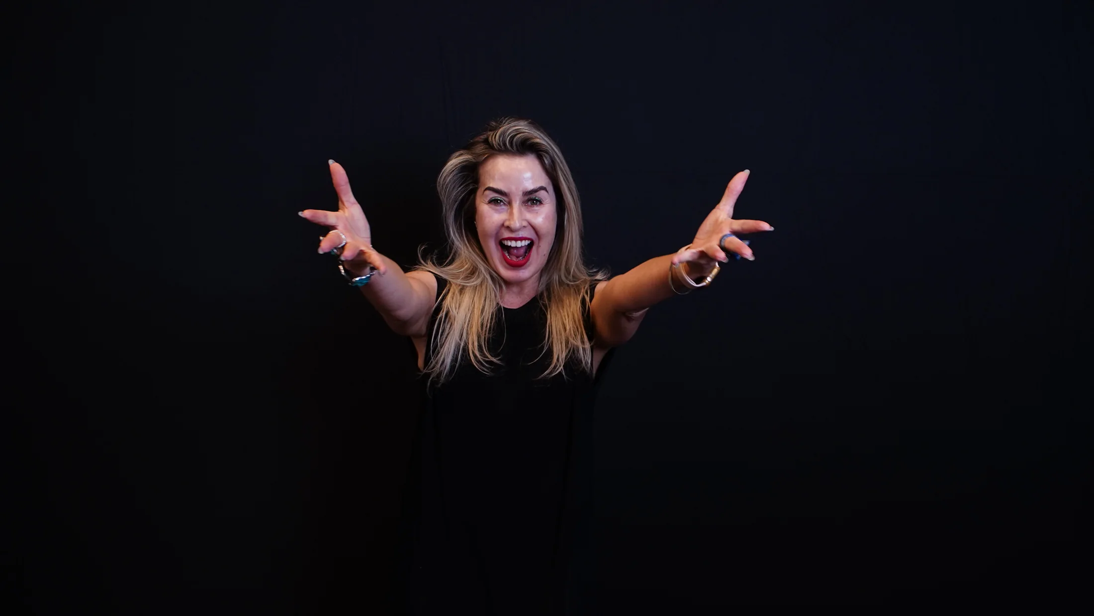 Daniella Prestes de braços abertos, rindo, em fundo escuro de estúdio
